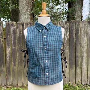 May include: A sleeveless button-down shirt featuring a green and navy blue plaid pattern. The garment includes a pointed collar, a front button placket, a single chest pocket, and black ribbon ties on both sides for an adjustable fit.