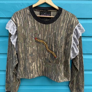 Op de afbeelding: Een shirt met lange mouwen met een camouflagepatroon en een decoratief duizendpotenontwerp. De shirt heeft een zwarte kraag en manchetten, met witte kant en oogjesdetails op de mouwen. De tekst "Devon In Space" is zichtbaar op de kraag.