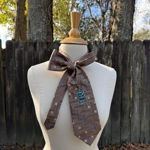 May include: A brown necktie with a geometric pattern of squares and rectangles in shades of brown, blue, and white. The tie features a beaded turquoise design. The tie is displayed on a mannequin.