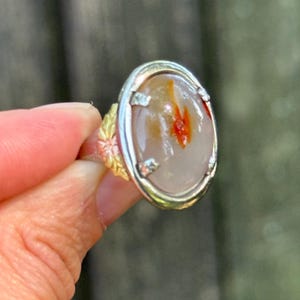 May include: A silver ring featuring an oval-shaped, translucent cabochon stone with a light gray base and a distinct reddish-orange inclusion in the center. The ring band is decorated with a textured, gold-toned leaf pattern on the sides.