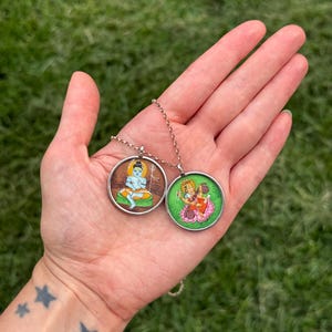 May include: Two round pendants on a silver chain, each featuring a colorful illustration. One pendant depicts a seated figure with blue skin, the other shows a figure on a lotus flower. The pendants are held against a green grassy background.