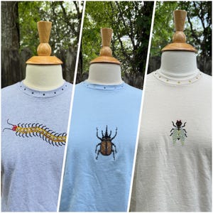 May include: Three t-shirts on display, each with a unique insect design. The shirts are light gray, light blue, and off-white. Each shirt has a beaded insect design and small studs around the neckline. The insects include a centipede, a beetle, and a fly.