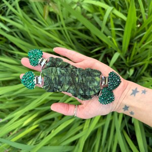 May include: A green and silver watch strap with a textured, dark green fabric centre. The strap features two decorative, sparkling green clusters on each end. The watch strap is shown against a background of green foliage.