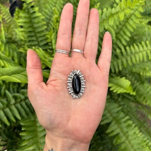 May include: An oval-shaped brooch featuring a central black stone set in a silver-toned metal frame. The frame is decorated with a series of small, raised silver spheres and thin, radiating metal lines that create a sunburst-like border pattern.