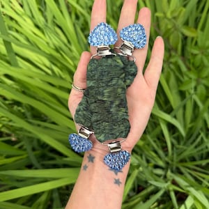 May include: Two green fabric hair clips with blue heart-shaped decorations. The clips have silver metal clasps. The heart-shaped decorations are covered in small blue crystals. The background is green foliage.