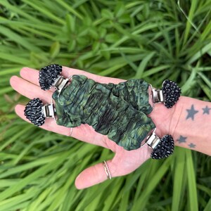 May include: A pair of green and black beaded bracelets with silver-tone clasps. The bracelets have a textured, crinkled fabric design in shades of green. The black beads are clustered at each end of the bracelet.