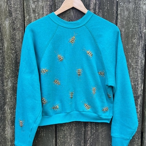 May include: A turquoise blue sweatshirt with a crew neck and long sleeves. The sweatshirt is decorated with a pattern of small, gold-coloured bugs.