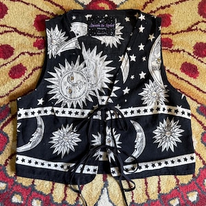 May include: A black sleeveless waistcoat featuring a white celestial print of suns, moons, and stars. The garment has a V-neckline and a front tie closure with black fabric strings. A label at the inner neckline reads Devon In Space by Oscar Hudson.