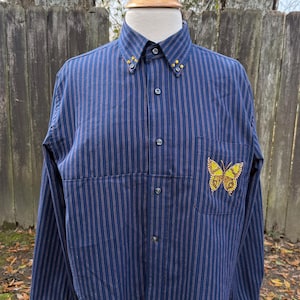 Op de afbeelding: Marineblauw overhemd met rode verticale strepen. Het overhemd heeft een gele, met kralen bezette vlinder op de zak en gele, met kralen bezette accenten op de kraag en manchetten. Het shirt met lange mouwen wordt op een mannequin gepresenteerd.