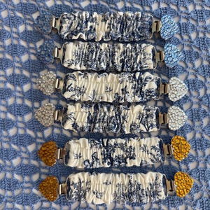 May include: Six elasticated fabric clips featuring a blue and white toile de Jouy pattern. Each clip has a metal fastener on both ends, topped with heart-shaped embellishments in either light blue, clear, or amber-coloured rhinestones.