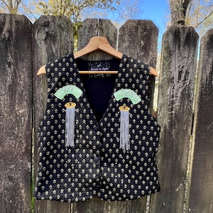 Op de afbeelding: Zwart vest met een gouden bloemenpatroon en decoratieve versieringen. Het vest heeft een V-hals en is aan de voorkant dichtgeknoopt. De versieringen omvatten groene waaiervormige details en gouden en zilveren kwastjes. De tekst "Devon In Space" is zichtbaar.