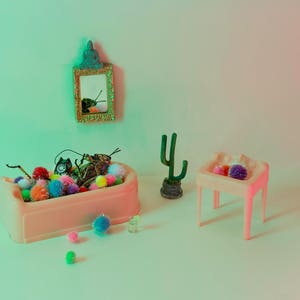 May include: A pink toy bath filled with colourful pom-poms and a toy bug. A small pink sink with a purple pom-pom is on the right. A green cactus and a gold framed mirror are in the background.