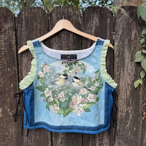 May include: A blue sleeveless crop top featuring a graphic of two chickadees perched on a branch surrounded by white blossoms. The garment has light green eyelet trim along the armholes, black side laces, and a label reading Devon In Space.