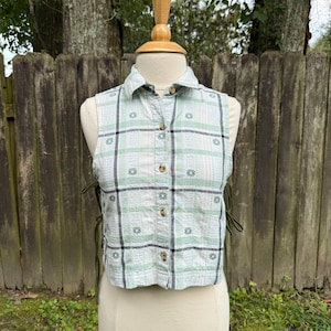 May include: A sleeveless, cropped button-down shirt featuring a light green, white, and black plaid pattern with small floral motifs. The garment includes a classic collar and dark green side laces that adjust the fit along the open sides.