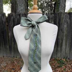 May include: A green and black patterned necktie with a bow, displayed on a mannequin. The tie features a central design of blue and gold beads. The bow and tie are adorned with small, sparkling embellishments.