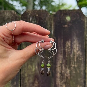 May include: A pair of silver earrings with a unique design. Each earring features a curved, openwork top with a green bead and a small, leaf-shaped charm. The earrings are held up against a blurred background.