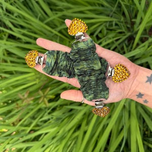 May include: Green fabric wrist cuffs with silver clasps and heart-shaped yellow rhinestone embellishments. The cuffs are shown against a background of green foliage.