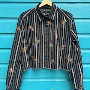 Op de afbeelding: Een zwart en grijs gestreept, getailleerd shirt met gouden insectenversieringen van kralen. Het shirt heeft lange mouwen en een knoopsluiting. De tekst "Dragon In Space" is zichtbaar op het label.