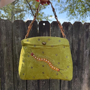 May include: A lime green handbag featuring a beaded centipede design in orange, yellow, and clear crystals. The bag is adorned with small scattered purple beaded flower motifs and includes a tan leather strap with metallic ring hardware connectors.