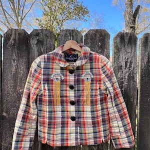 May include: A plaid jacket with a black, red, and white pattern. The jacket has two gold eye-shaped embellishments with fringe on the front. The label on the jacket reads "Dream In Space".