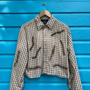 Op de afbeelding: Een bruin en beige geruit overhemd met lange mouwen en een knoopsluiting. Het overhemd heeft een verkorte lengte en beschikt over ingewikkelde parelversieringen in een geometrisch patroon.