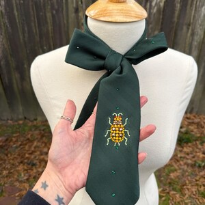 May include: A dark green necktie with a bow at the top, adorned with small, sparkling green and yellow embellishments. The tie features a detailed insect design in the center, crafted with yellow and orange beads. The tie is displayed on a mannequin.