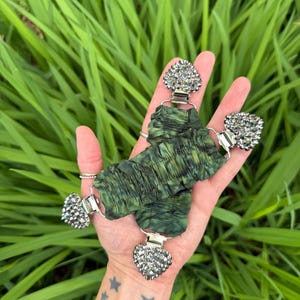May include: A green fabric accessory with silver-toned heart-shaped clasps adorned with sparkling rhinestones. The fabric has a textured, crinkled appearance. The accessory is displayed against a backdrop of green foliage.