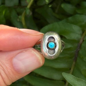 May include: A silver ring featuring a smooth, oval-shaped centrepiece with a recessed dark area surrounding a small, round, bright blue turquoise stone set in the middle. The metal band is polished and reflects light on its rounded surface.
