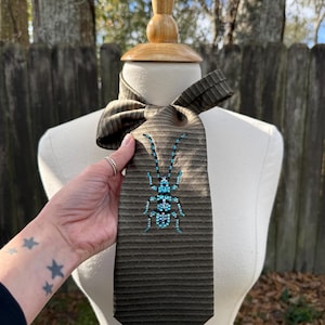 May include: A brown and black striped tie with a beaded turquoise insect design. The tie is displayed on a mannequin. The tie is held by a hand with star tattoos.