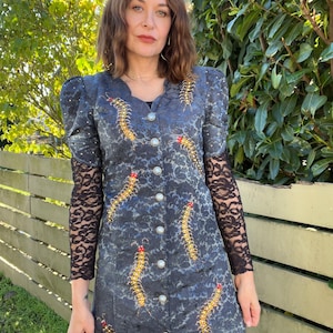 May include: A blue dress with a long-sleeved black lace overlay. The dress is decorated with gold and red beaded centipedes.