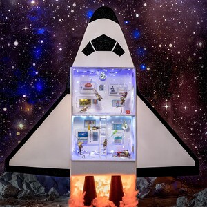 May include: A whimsical, multi-level space shuttle diorama with miniature figures and accessories inside. The shuttle is white with black accents, set against a starry background. The bottom section features illuminated orange flames, suggesting a launch. The interior is lit with small lights.