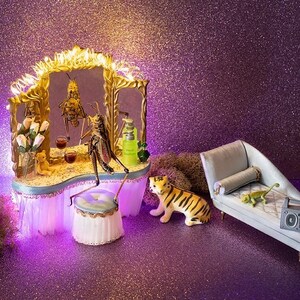 May include: A miniature bar scene with a gold-framed mirror, a grasshopper figure, and drinks. A tiger figurine and a chaise lounge with a boombox are also present. The background is a glittery purple, creating a whimsical and playful atmosphere.