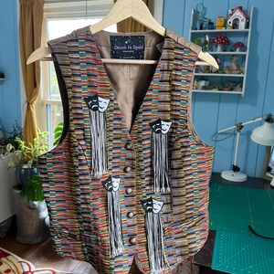 May include: A brown vest with a multi-colored striped pattern. The vest has four black and white theater masks with tassels attached. The vest has a label that says "Devon in Space".