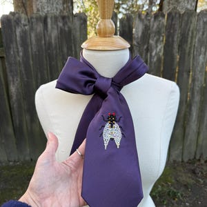 May include: A deep purple necktie with a decorative fly design. The fly is embellished with black, red, and white beads. The tie is displayed on a mannequin.