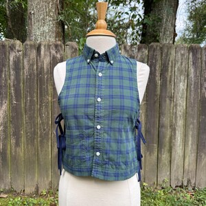 May include: A sleeveless button-down shirt featuring a green and navy blue plaid pattern. The garment has a pointed collar, a front button placket, and dark blue ribbon ties on both sides. A single patch pocket is visible on the lower front.