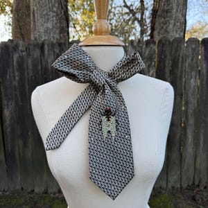 May include: A patterned necktie with a bow, displayed on a mannequin. The tie is primarily gray with a small, repeating pattern. A decorative brooch with red and clear accents adorns the tie. The background features a wooden fence and trees.