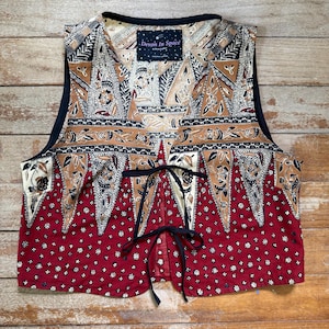 May include: A sleeveless waistcoat featuring a dark red lower section with a small white floral pattern and an upper section with tan, black, and white geometric and floral motifs. The garment has a black trim, a V-neckline, and two black tie closures.