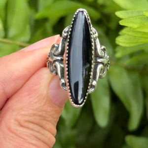 May include: A silver ring featuring a long, narrow, marquise-shaped black stone set in a beaded metal bezel. The band displays ornate, scrolling metalwork on both sides of the central stone, creating a detailed and structured appearance.