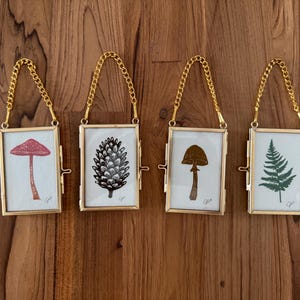 May include: Four gold-toned picture frames with chains hanging from a wooden surface. Each frame contains a black and white illustration of a mushroom, a pine cone, a fern, and a mushroom with a red cap.