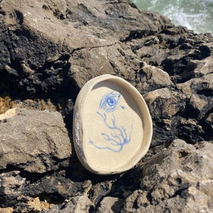 Puede incluir: Un pequeño plato de cerámica blanquecina con un diseño de ojo y ramas azules. El plato, de forma ovalada, está colocado sobre una superficie rocosa. El diseño pintado a mano añade un toque único. El plato se utiliza probablemente para guardar objetos pequeños.