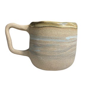 Puede incluir: Una taza de cerámica hecha a mano con un asa curva única. La taza presenta un exterior beige texturizado con sutiles rayas horizontales azules y marrones. El borde tiene un acabado más oscuro y vidriado, agregando contraste. Ideal para café o té.