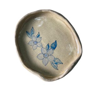 Puede incluir: Un plato de cerámica hecho a mano con una superficie beige moteada. Dos diseños florales azul claro con hojas azules están pintados en el plato. El plato tiene una forma irregular y orgánica y un borde elevado.
