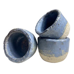 Puede incluir: Conjunto de cuatro pequeñas tazas de cerámica hechas a mano. Las tazas son de un azul apagado con una superficie texturizada y una base beige natural sin esmaltar. Las tazas están apiladas.