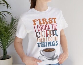 May include: White t-shirt with the text "First I drink the coffee then I do the things" in colorful letters. A cup of coffee is pictured below the text.
