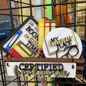 May include: A wooden sign with the text "Certified Bookworm" in gold letters. There are other signs with book-themed designs, including "So much to read, so little time" and "My happy place".