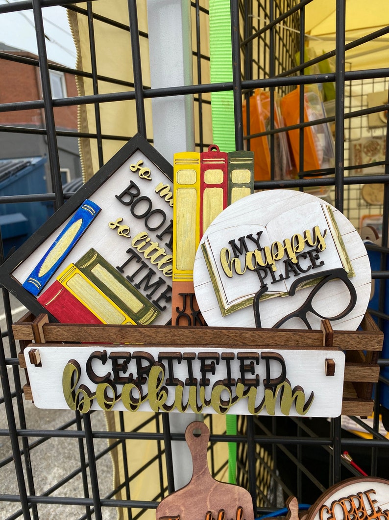 May include: A wooden sign with the text "Certified Bookworm" in gold letters. There are other signs with book-themed designs, including "So much to read, so little time" and "My happy place".
