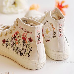 May include: Cream-colored high-top sneakers with floral embroidery in red, orange, and green. The sneakers feature gold eyelets, and the text "Mrs. Krass" and a date are embroidered on the side. The Converse All Star logo is visible.