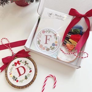 Puede incluir: Un kit de bordado con temática navideña con un adorno de aro de madera con la letra "D" y un lazo rojo. El kit incluye hilo de bordar, un parche redondo con la letra "F" y un bastón de caramelo.