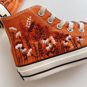 May include: Close-up of an orange high-top canvas shoe with embroidered floral design. The embroidery features white, orange, and brown flowers and foliage. The shoe has white laces, eyelets, and a white rubber sole with black stripes.