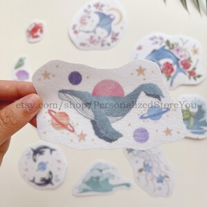 May include: A collection of fabric stickers featuring celestial and marine themes. The central sticker depicts a blue whale with a pink sun, surrounded by planets and stars. Other stickers show dolphins, stingrays, and floral elements, all in soft pastel colors.